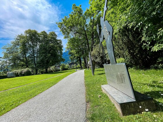 Stadtrunde mit Metallskulptur Ein geschotterter Spazierweg führt an einer Metallskulptur und grünen Wiesen mit Bäumen vorbei.