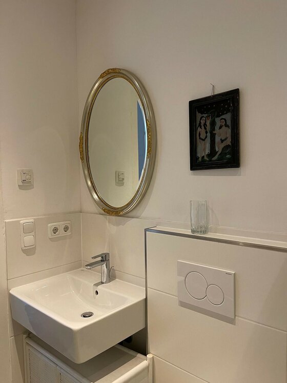 Badezimmer mit weißem Waschbecken, ovalem Spiegel mit Goldrahmen und einem kleinen Wandbild.