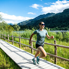 Eine sportliche Frau joggt auf einem Holzsteg entlang eines malerischen Sees, umgeben von grünen Wiesen und Bergen.