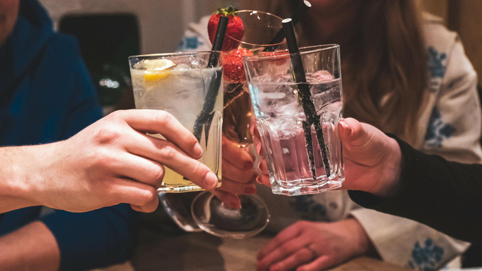 Drei Hände stoßen mit bunten Cocktails an, während im Hintergrund fröhliche Gesichter zu sehen sind, die den Moment genießen.