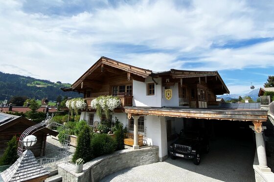 Villa Kitz im Sommer Alpines Wohnhaus aus Holz und Stein mit Blumenbalkonen und Carport vor Bergkulisse.