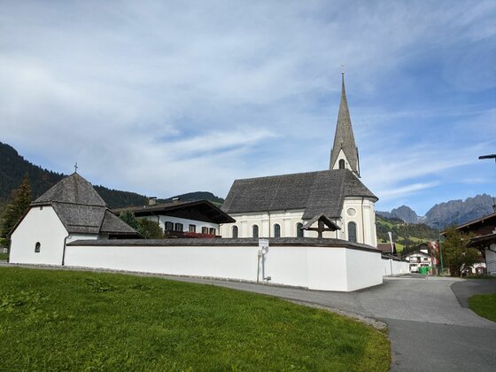 Weiße Kirche mit spitzem Turm und umlaufender Mauer vor bewaldeten Bergen.