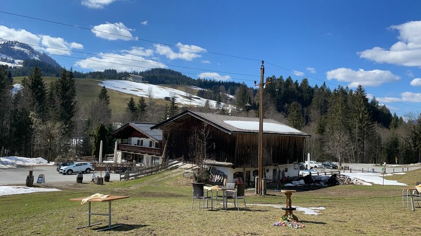 Burgeralm Spielbergalm Alpines Holzgebäude mit Parkplatz und Außenbestuhlung vor bewaldeten Bergen mit Schneeresten.