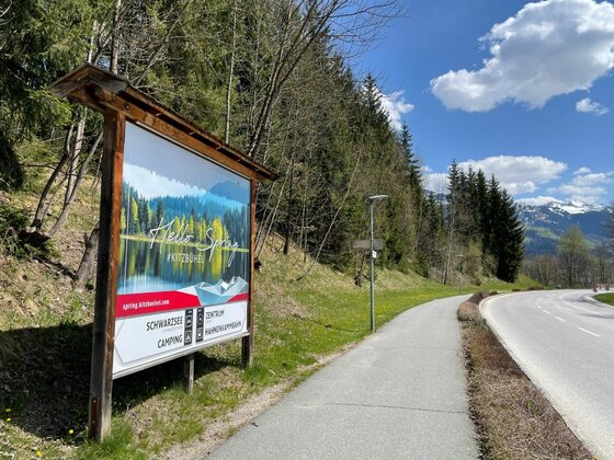 Infotafel am Radweg Große Informationstafel an einem asphaltierten Rad- und Gehweg neben einer Landstraße vor bewaldetem Hang.