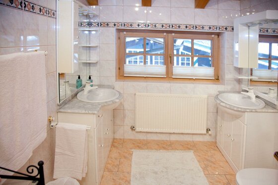 Egger Studio Hornblick Badezimmer Badezimmer mit zwei Waschbecken, hellen Fliesen und einem Holzfenster.