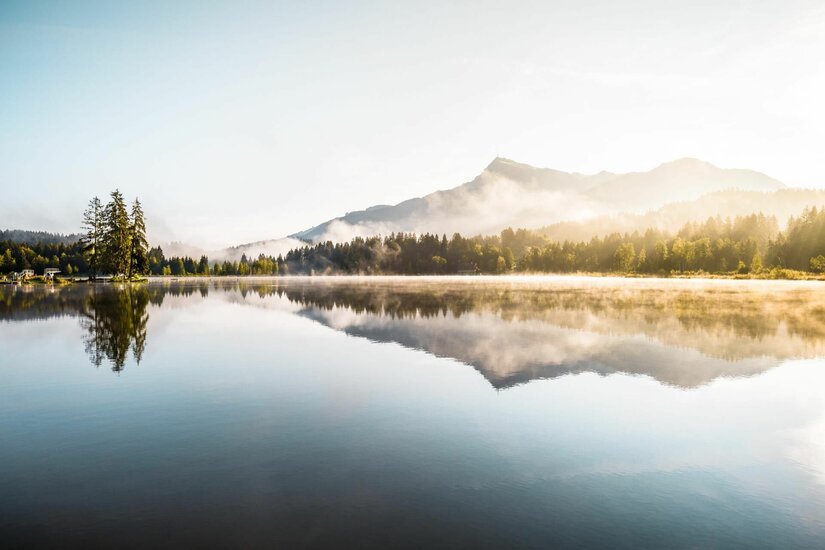 Schwarzsee bei Kitzbühel Ein ruhiger See mit Spiegelung der Berge und Nadelbäume bei leichtem Morgennebel.