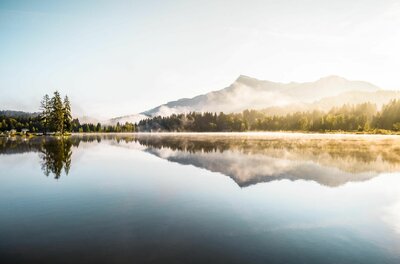 Ein ruhiger See mit Spiegelung der Berge und Nadelbäume bei leichtem Morgennebel.