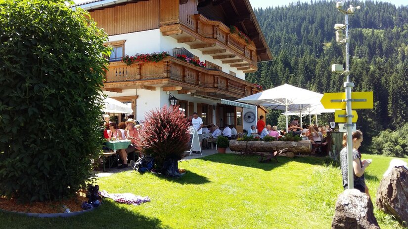 Going_Hollenauer Kreuz_außen_Sommer_2022_2 Ein gemütliches Restaurant mit Sonnenschirmen und Gästen im Freien, umgeben von grünen Wiesen und Bergen. Die Fassade des Hauses ist im traditionellen Stil mit Blumenkästen dekoriert.