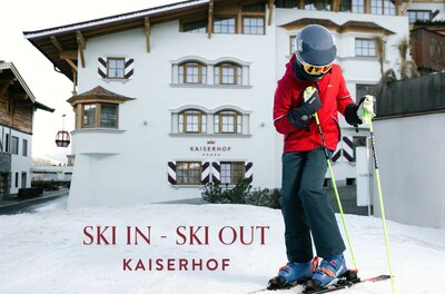 Hotel-Kaiserhof-Kitzbühel-Ski-In-Ski-Out Ein Skifahrer steht auf Schnee vor dem Hotel Kaiserhof. Im Hintergrund sind eine traditionelle Alpenarchitektur und der Schriftzug „SKI IN - SKI OUT“ zu sehen.