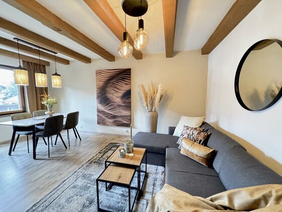 Ein modernes Wohnzimmer mit einer gemütlichen Couch, einem Kaffee­tisch und einem Esstisch. Große Fenster und stilvolle Dekoration schaffen eine einladende Atmosphäre.