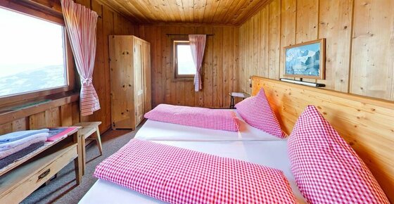 Zimmer der Hocheckhütte Rustikales Doppelzimmer mit Holzverkleidung, karierter Bettwäsche und Fenster mit Bergblick.