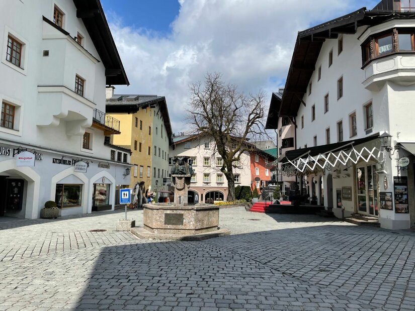 Dorfplatz mit Brunnen Gepflasterter Dorfplatz mit einem Steinbrunnen im Zentrum, umgeben von mehrstöckigen Häusern mit hellen Fassaden.