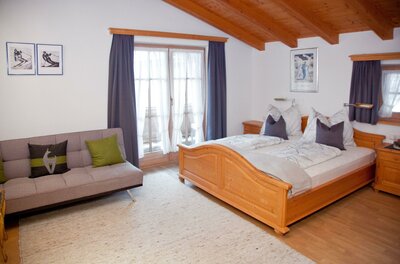 Egger Studio Hornblick Schlafzimmer mit Doppelbett aus Holz, grauem Sofa und Balkontür unter einer Holzdecke.