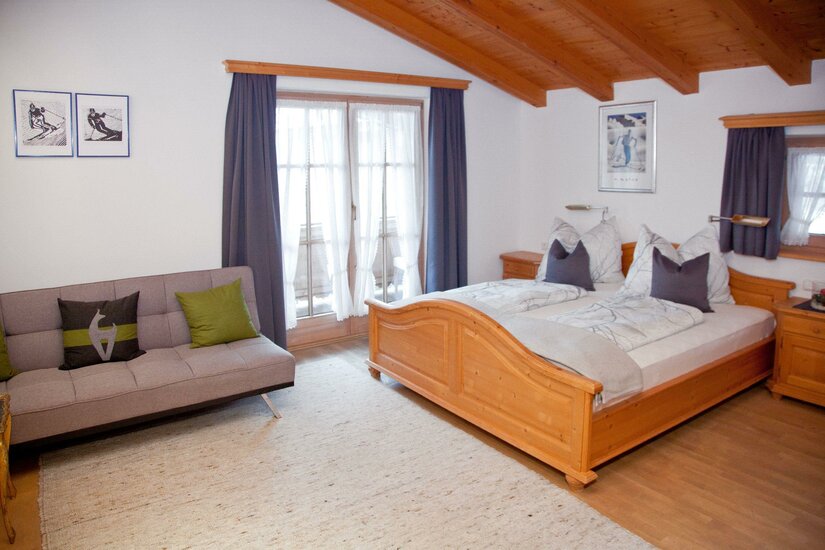 Egger Studio Hornblick Schlafzimmer mit Doppelbett aus Holz, grauem Sofa und Balkontür unter einer Holzdecke.