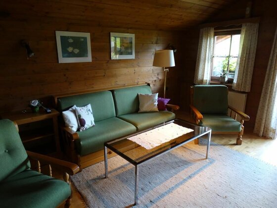 Wohnraum im Haus Lindler Rustikaler Wohnraum mit grünem Sofa, Sesseln und Glastisch vor holzverkleideten Wänden.