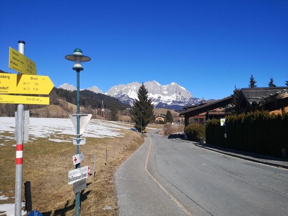 Wanderwegweiser an der Astberg-Runde Gelbe Wanderwegweiser an einer asphaltierten Straße mit Blick auf schneebedeckte Berge unter blauem Himmel.