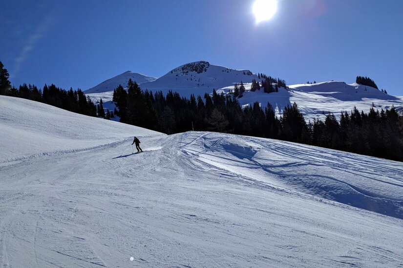 Ein Skifahrer auf einer präparierten Piste vor verschneiten Berggipfeln und blauem Himmel bei Sonnenschein.
