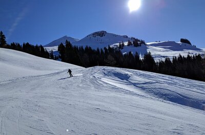 Ein Skifahrer auf einer präparierten Piste vor verschneiten Berggipfeln und blauem Himmel bei Sonnenschein.