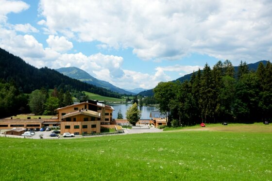Ums Bichlach am See Blick über eine grüne Wiese auf ein Holzgebäude vor einem See und bewaldeten Bergen.