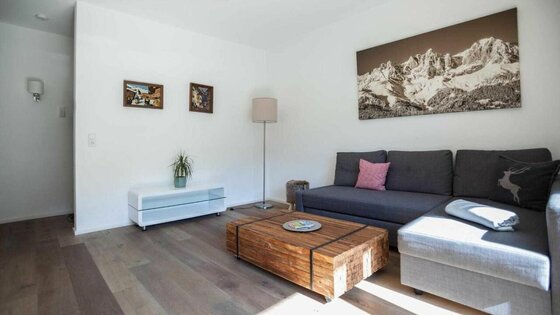 Wohnraum im Kitz Apartment Modernes Wohnzimmer mit grauem Ecksofa, Holztisch und großem Bergbild an der weißen Wand.