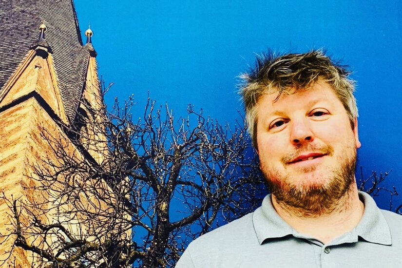 Akupunkturpraxis Kitzbühel Ein Mann mit Bart vor einem Kirchturm und einem kahlen Baum unter blauem Himmel.