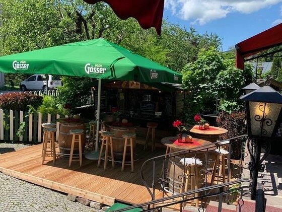 Außenbereich einer Bar mit Holzterrasse, Weinfässern als Stehtische und grünem Sonnenschirm.