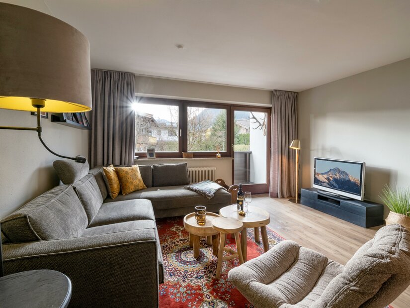 Modernes Wohnzimmer mit grauem Ecksofa, Holztischen, Orientteppich und Fernseher vor einem Fenster mit Bergblick.