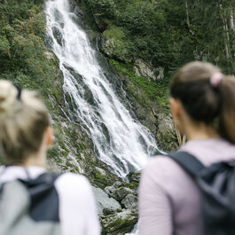 Der Sintersbacher Wasserfall stürzt über grüne Felsen, während zwei Wanderer fasziniert auf das Naturschauspiel blicken.