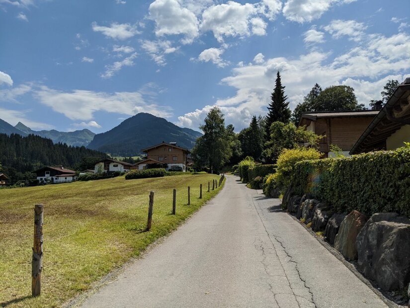 Asphaltierter Weg führt an einer Wiese und Häusern vorbei auf bewaldete Berge unter bewölktem Himmel zu.