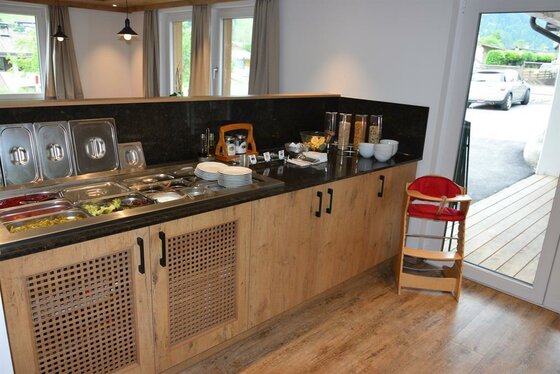 Buffetbereich mit Speisebehältern aus Edelstahl auf einer Holztheke und ein Kinderhochstuhl daneben.
