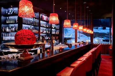 Rubin Bar im Hotel Moderne Hotelbar mit roten Hängelampen, Barhockern und Blick auf eine verschneite Winterlandschaft.