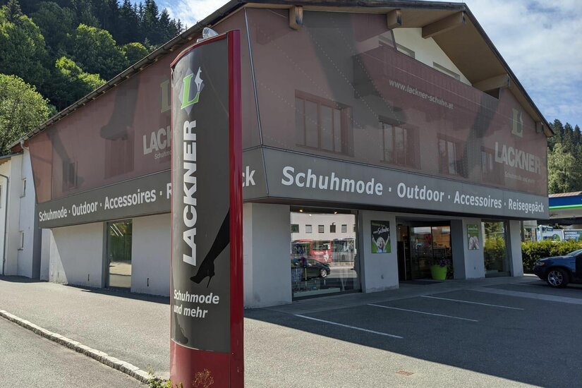 Lackner Schuhe Fachgeschäft Außenansicht des Schuhgeschäfts Lackner mit Werbestele und Parkplätzen vor dem Gebäude.