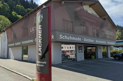 Außenansicht des Schuhgeschäfts Lackner mit Werbestele und Parkplätzen vor dem Gebäude.