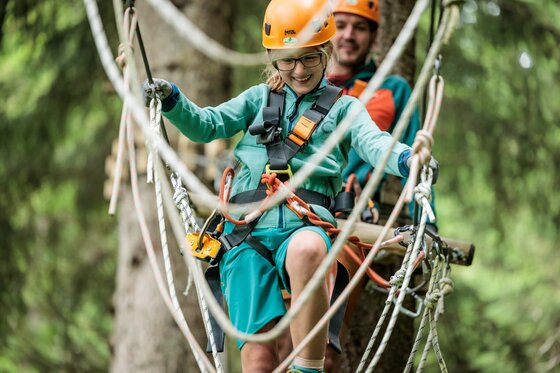 Ein Kind und ein Erwachsener mit Helmen und Klettergurten balancieren auf Seilen in einem Hochseilgarten.