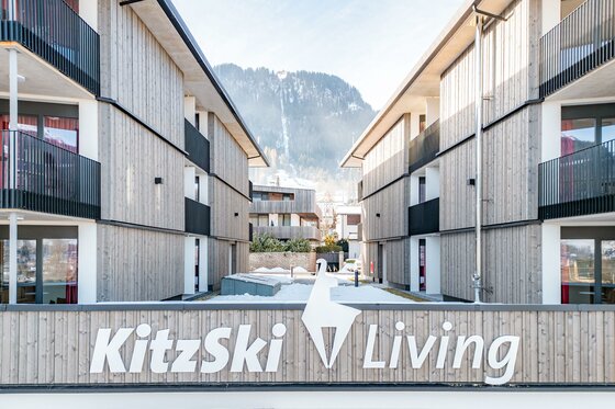 Moderne Holzbauten mit Balkonen, umgeben von Bergen. Das Schild "KitzSki Living" steht im Vordergrund.