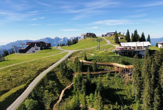 Sonnenrast Trail bei Bergstation Blick auf Seilbahnstationen, eine Berghütte und einen hölzernen Mountainbike-Trail im alpinen Gelände.