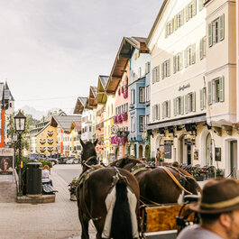 Eine malerische Straße in Kitzbühel, gesäumt von bunten Gebäuden und blühenden Pflanzen, mit Pferden im Vordergrund.