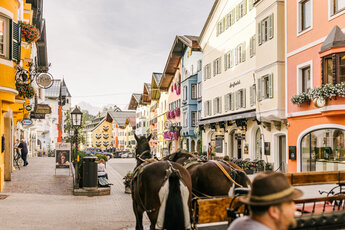 Eine malerische Straße in Kitzbühel, gesäumt von bunten Gebäuden und blühenden Pflanzen, mit Pferden im Vordergrund.