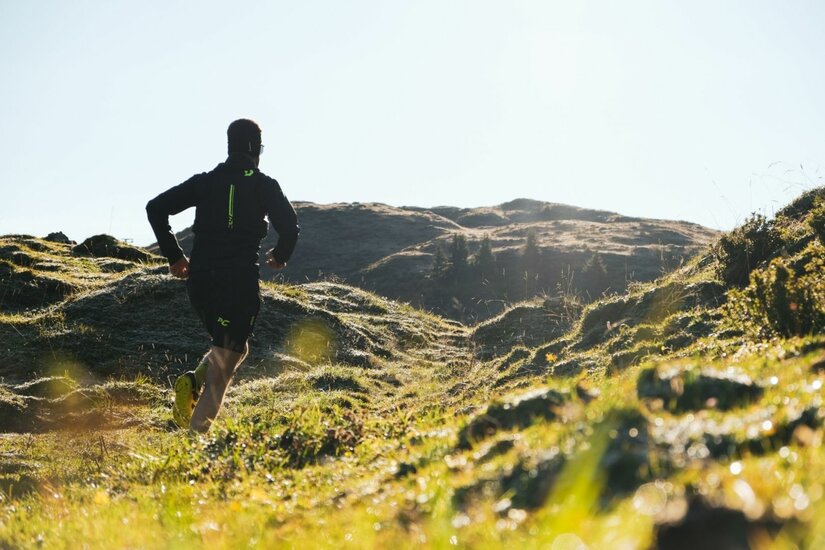 Trailrunning mit Paul Doyle Ein Trailrunner läuft von hinten gesehen über eine hügelige, grüne Almwiese in Richtung eines Bergrückens.