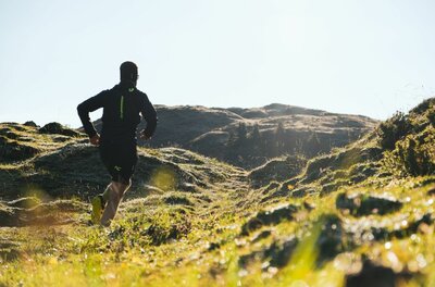 Trailrunning mit Paul Doyle Ein Trailrunner läuft von hinten gesehen über eine hügelige, grüne Almwiese in Richtung eines Bergrückens.