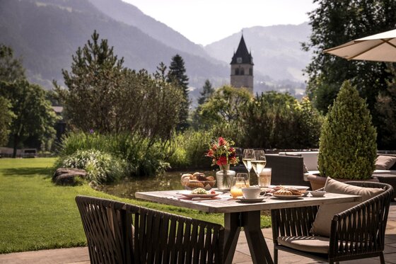 Champagnerfrühstück auf der Terrasse Hotel Kitzhof Ein schöner Garten mit einem Tisch, auf dem Frühstück und Getränke serviert werden. Im Hintergrund sind Berge und ein Kirchturm sichtbar.