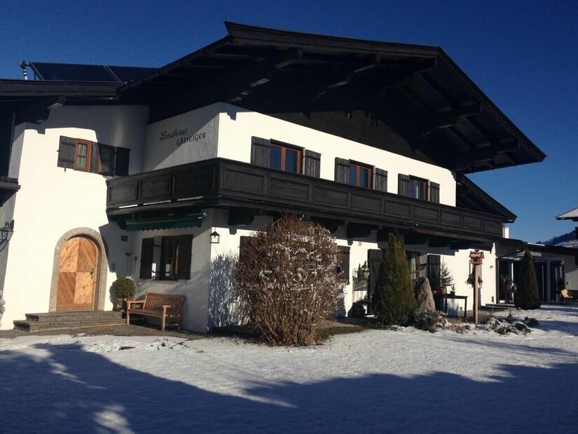 Landhaus Gasteiger im Winter Zweistöckiges weißes Landhaus mit dunklem Holzbalkon und Schneefläche im Vordergrund bei blauem Himmel.
