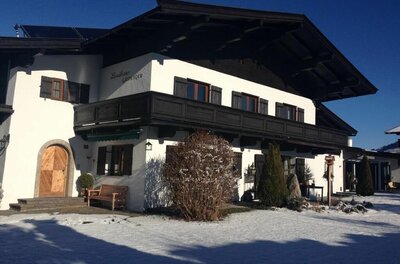 Landhaus Gasteiger im Winter Zweistöckiges weißes Landhaus mit dunklem Holzbalkon und Schneefläche im Vordergrund bei blauem Himmel.