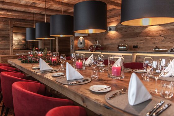 Speisebereich der Hahnenkamm Lodge Gedeckter Holztisch mit roten Sesseln, weißen Servietten und schwarzen Hängelampen in einer Lodge.