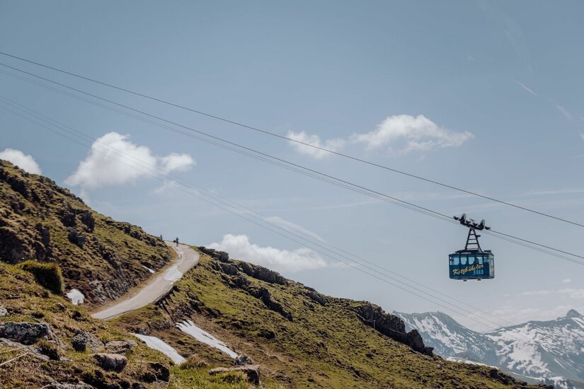 Seilbahn am Kitzbüheler Horn Eine blaue Seilbahnkabine schwebt vor einem Berghang mit Wanderweg und schneebedeckten Gipfeln im Hintergrund.