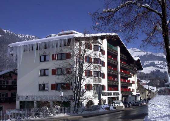 Q! Hotel Maria Theresia Mehrstöckiges Hotelgebäude mit Schneelast und Eiszapfen am Dachrand vor verschneiten Bergen.