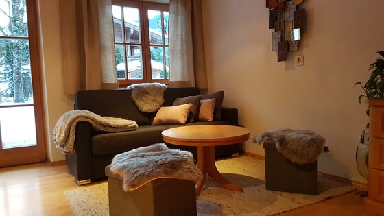 Wohnbereich mit dunklem Sofa, rundem Holztisch und zwei Hockern mit Fellauflage vor einem Fenster.