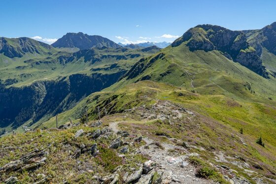 Wanderweg am Gebirgskamm Ein schmaler Wanderpfad führt über einen grasbewachsenen Gebirgskamm mit Blick auf umliegende Berggipfel.