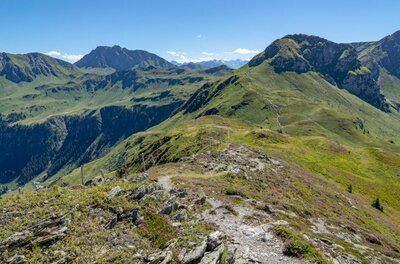 Wanderweg am Gebirgskamm Ein schmaler Wanderpfad führt über einen grasbewachsenen Gebirgskamm mit Blick auf umliegende Berggipfel.