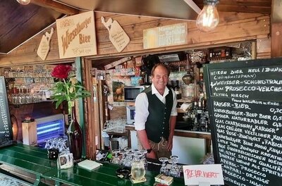 Fritz' LICHTBLICK Ausschank Ein Mann in Trachtenweste steht hinter einem hölzernen Tresen mit Getränkekarte und Gläsern.